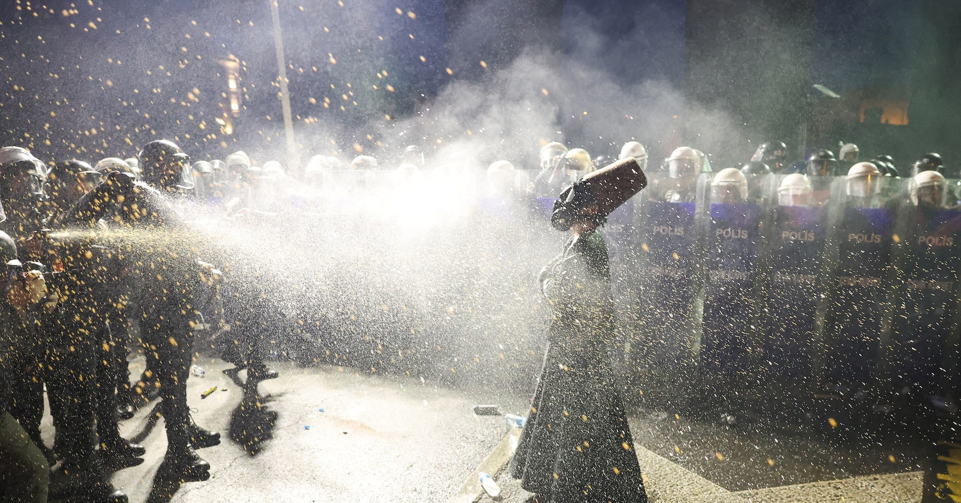 A person in a black robe and a tall brown felt hat stands facing a line of riot police holding shields labeled “Polis.” The person is being sprayed with pepper spray, and the air is filled with mist and debris. The person wears a gas mask and stands with a composed posture amid the chaos. A person in a black robe and a tall brown felt hat stands facing a line of riot police holding shields labeled “Polis.” The person is being sprayed with pepper spray, and the air is filled with mist and debris. The person wears a gas mask and stands with a composed posture amid the chaos.